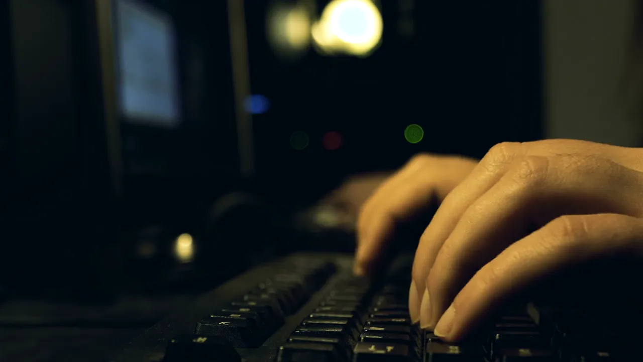 Mãos digitando em um teclado diante de monitores, em sala escura